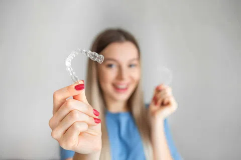 Smiling woman holding clear dental aligners for teeth straightening in a bright, neutral setting.
