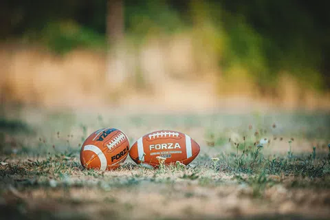 Two brown FORZA American footballs resting on dry grass with a blurred green background in daylight.