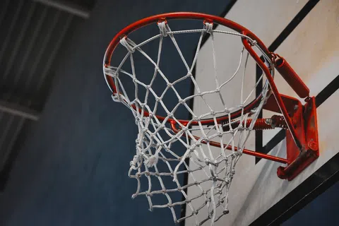 Close-up of a red basketball hoop with white net against a gray and blue indoor gym background.