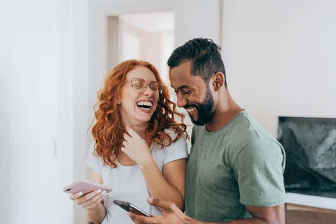 Couple laughing together while holding smartphones in a cozy modern living room setting