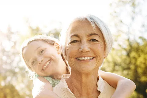 Smiling elderly woman with short white hair carrying a happy young girl on her back outdoors in a sunlit park