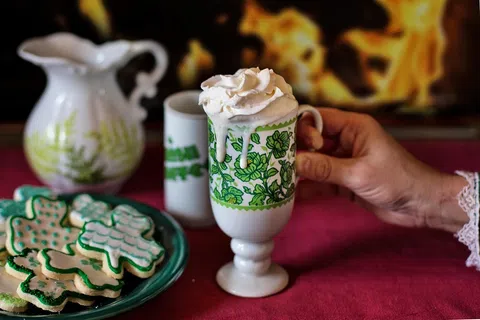 Hand holding vintage green floral mug with whipped cream hot drink, holiday cookies and fireplace in background.