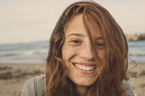 Smiling young woman with red hair and freckles wearing a knitted sweater at the beach on a cloudy day