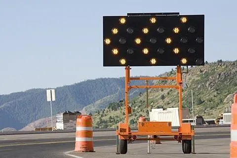 Electronic arrow sign directing traffic right on road near orange construction barrels with mountainous background