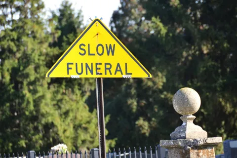 Yellow triangular warning sign reading Slow Funeral near cemetery stone and trees in background