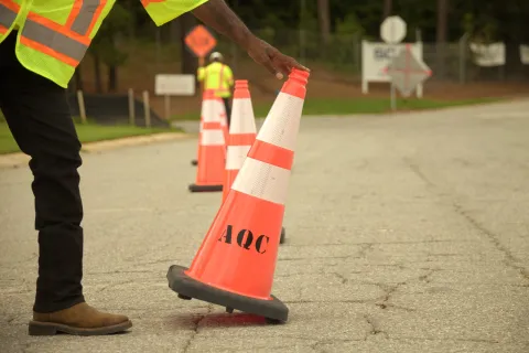 Worker in safety vest placing orange traffic cone with AQC on cracked asphalt road in daylight.
