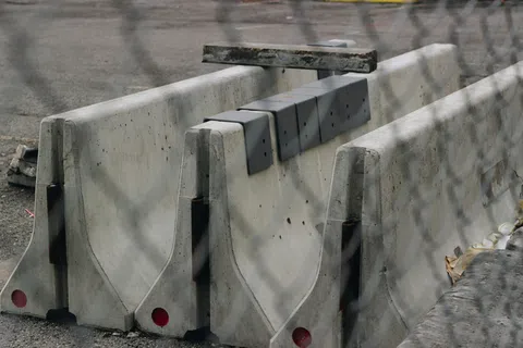 Stacked concrete road barriers with red reflectors behind a chain-link fence at a construction site.