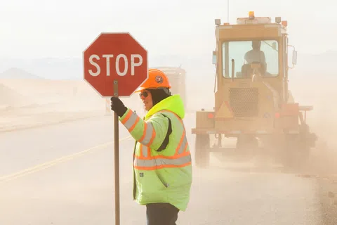 Construction worker in reflective jacket holding stop sign on dusty road with heavy machinery behind.