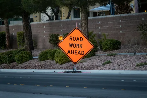 Orange diamond-shaped road sign reading 'Road Work Ahead' placed near street with trees and bushes behind.