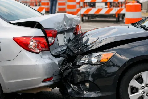 Two cars collide in a rear-end accident at a road work zone with orange cones and warning signs.