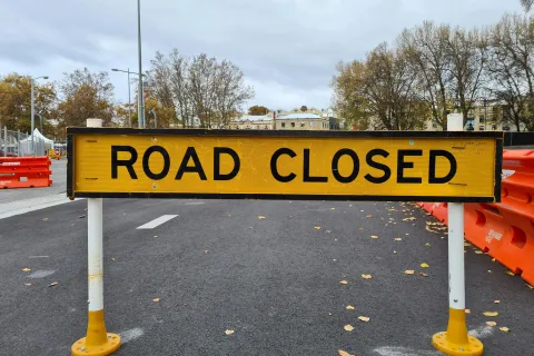 Yellow road closed sign blocking street with orange barriers and trees in the background on a cloudy day