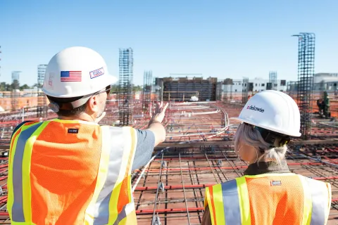 Two construction workers in safety gear inspect rebar framework at a sunny building site under clear blue sky.
