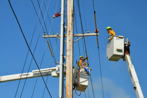 Two linemen in bucket trucks working on a wooden utility pole against a clear blue sky.