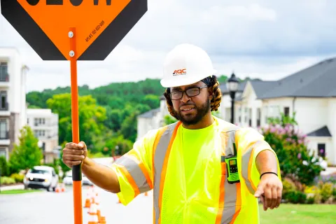 Construction worker wearing safety gear holding slow sign on suburban street with houses and greenery in background