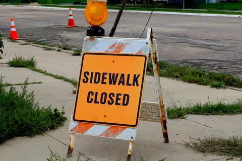 Orange sidewalk closed sign with warning light on a cracked urban sidewalk near traffic cones.
