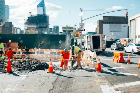Construction workers repairing urban road with safety cones, barriers, and city buildings in the background.