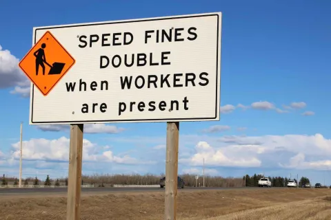 Road sign warning that speed fines double when workers are present, with blue sky and highway in background.