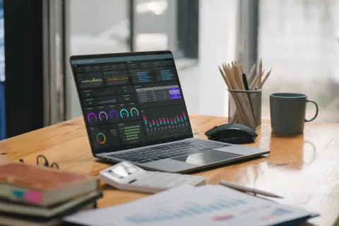 Open laptop displaying colorful business analytics dashboards on a wooden desk with office supplies and coffee cup.