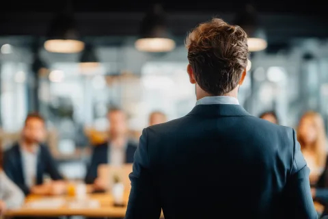Businessman in suit addressing a meeting with colleagues seated around a conference table in a modern office.