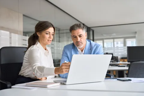 Two business professionals discussing work while looking at a laptop in a modern office setting