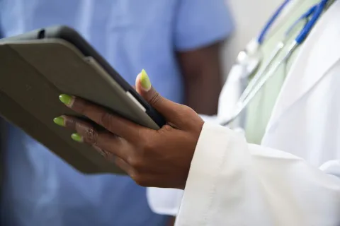 Close-up of a healthcare professional with green nails holding a tablet, wearing a white coat and stethoscope.