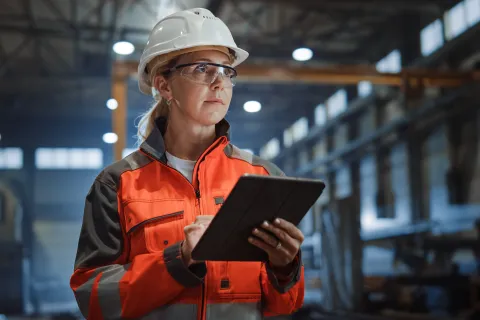 Female engineer wearing safety gear using tablet inside industrial warehouse with machinery and equipment.