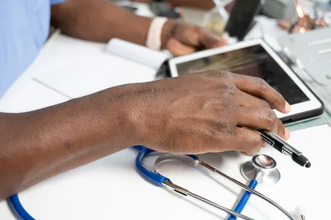 Close-up of a doctor's hand using a tablet with a blue stethoscope on a white desk.