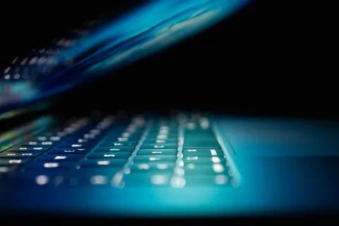 Close-up of a laptop keyboard with glowing backlit keys reflecting on the screen in low light.