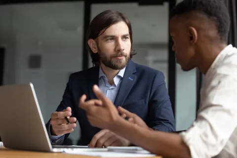 Two business professionals engaged in a serious discussion in a modern office setting with a laptop open.