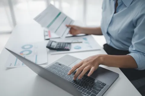 Person working on laptop with financial documents and calculator on desk in bright office setting