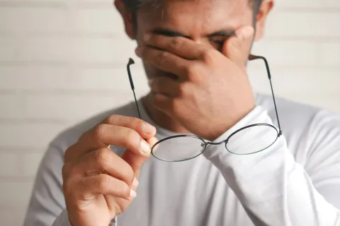 Man in white shirt holding glasses and rubbing eyes, showing signs of tiredness or eye strain indoors.