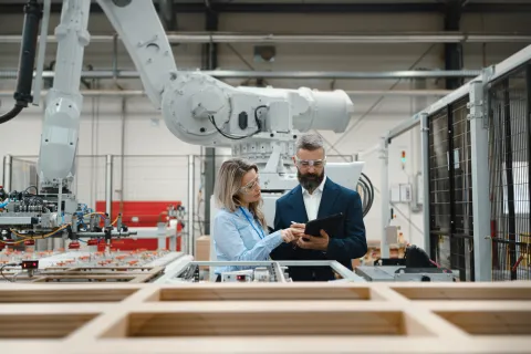 Two engineers in safety glasses discussing work on a tablet inside a modern automated manufacturing facility.