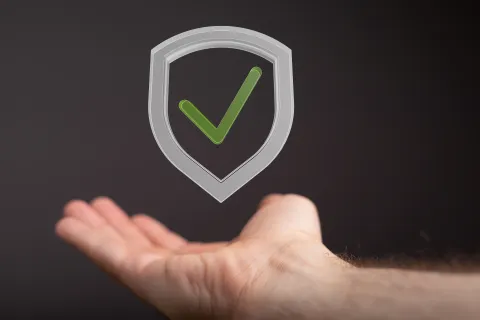 Open hand beneath a floating gray shield with a green checkmark symbolizing security and trust.