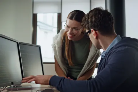 Two coworkers discuss coding on dual monitors in a bright modern office space.