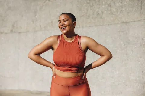 Smiling woman in orange sportswear standing with hands on hips against a textured concrete wall