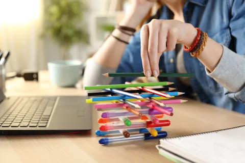 A person stacking colorful pens and pencils on a desk near a laptop, showcasing creativity in workspace.