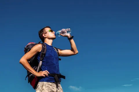 Young male hiker wearing backpack drinks water from bottle under clear blue sky outdoors
