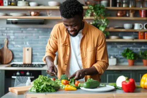 Man in yellow shirt chopping fresh vegetables in a modern kitchen surrounded by healthy produce and plants, highlighting the benefits of a nutritional diet.