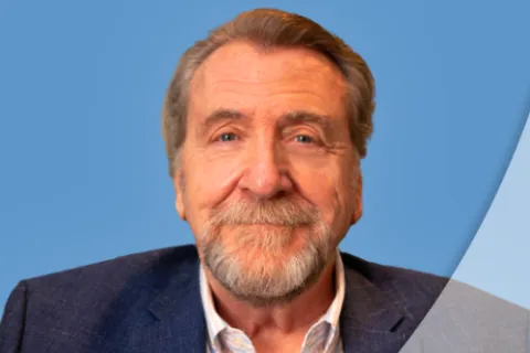 Smiling older man with gray beard wearing a blue blazer and striped shirt on a blue background.