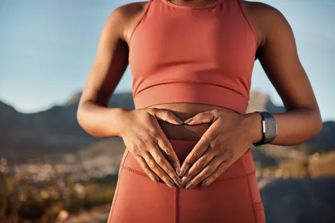 Woman in rust-colored sportswear forming a heart shape with hands over her stomach outdoors.
