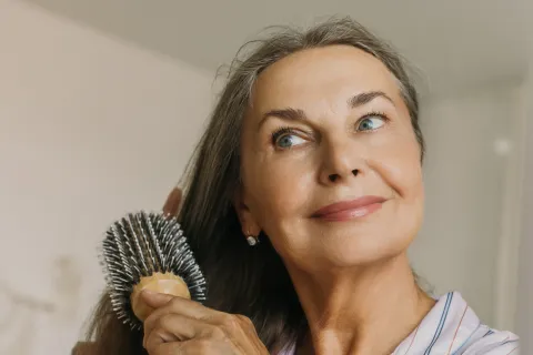 Older woman in striped pajamas brushing long gray hair with a round hairbrush, smiling gently.