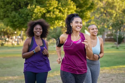 Three women jogging happily in a park wearing athletic wear and fitness trackers on a sunny day