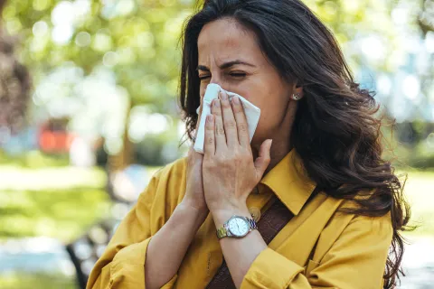 Woman sneezing outdoors during springtime surrounded by blooming flowers and pollen, representing seasonal allergy symptoms
