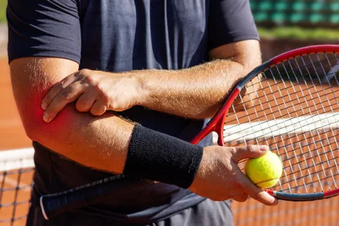 Tennis player with racquet and ball holding elbow showing pain or injury on clay court.