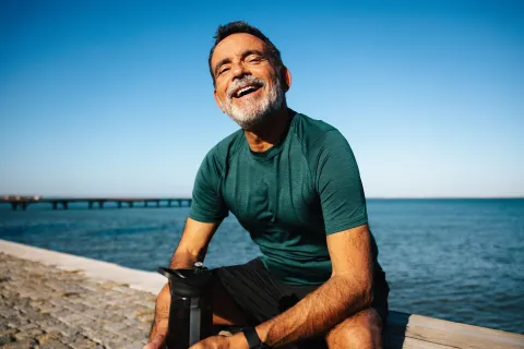 Smiling middle-aged man in sportswear sitting by the water with a fitness shaker bottle on a sunny day.
