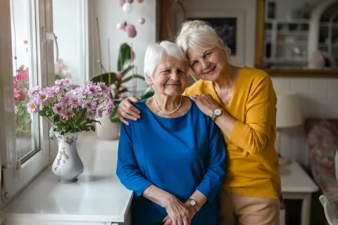 Two smiling women embrace in a cozy room with flowers, exuding warmth and togetherness.