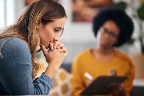 Worried woman sitting with hands clasped while therapist takes notes in counseling session.