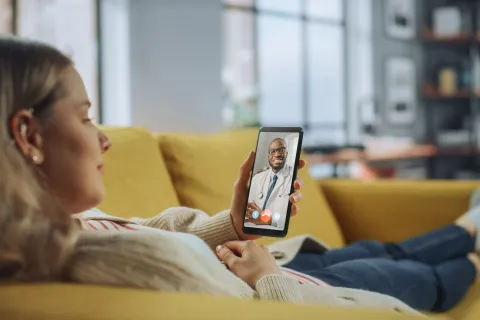 Woman having a video call with her primary care doctor on a smartphone while lying on a yellow couch at home