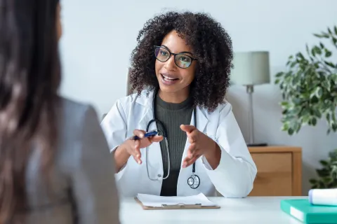 Female doctor with glasses and curly hair explaining something to a patient in a bright office setting.