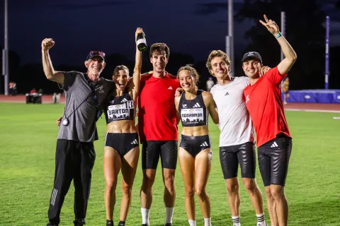 Group of six athletes celebrating on a track field at night, smiling and raising arms in victory.